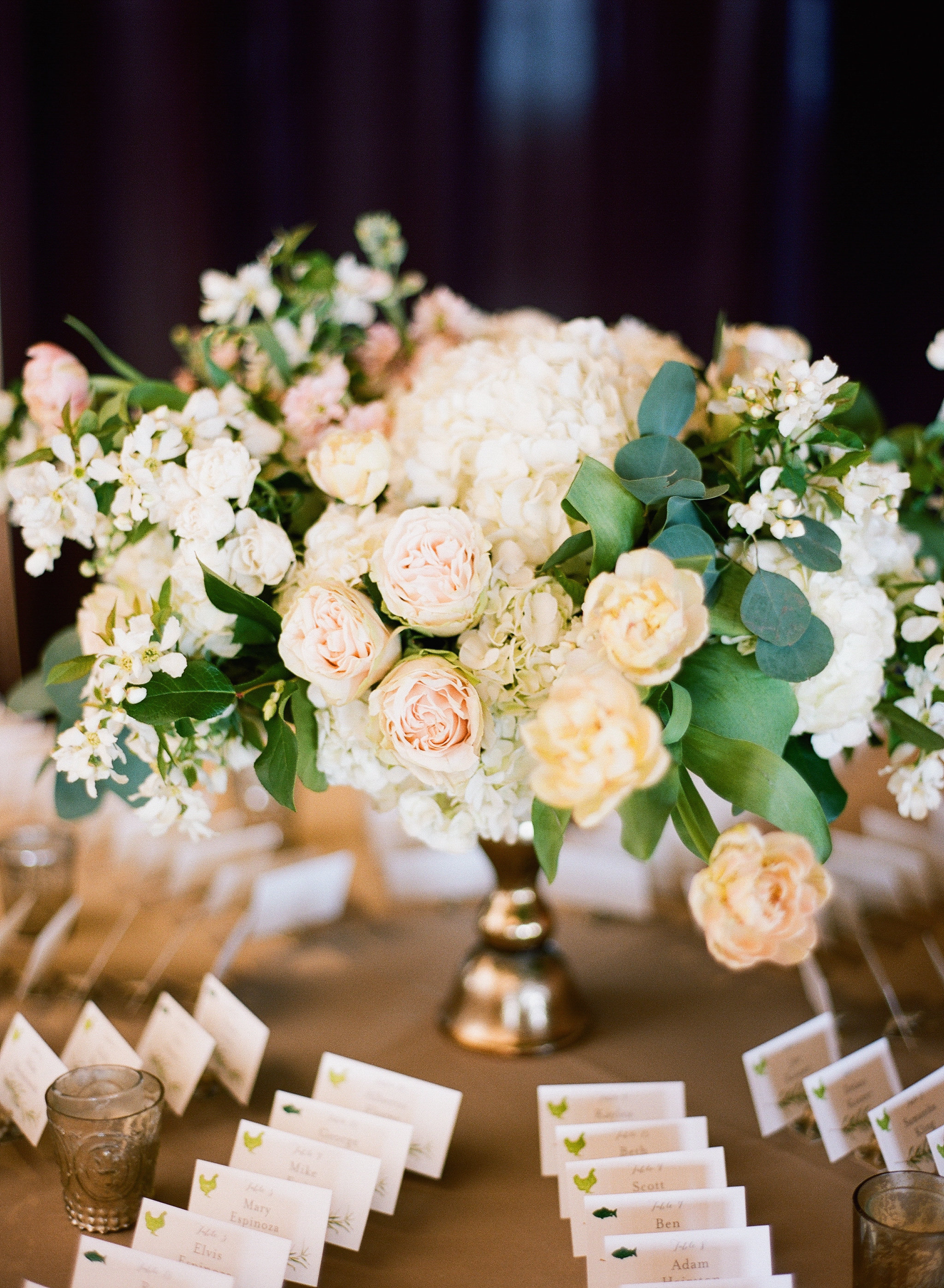 Escort card table flower arrangement for spring wedding with flowering branches, garden roses, tulips in gold vase.