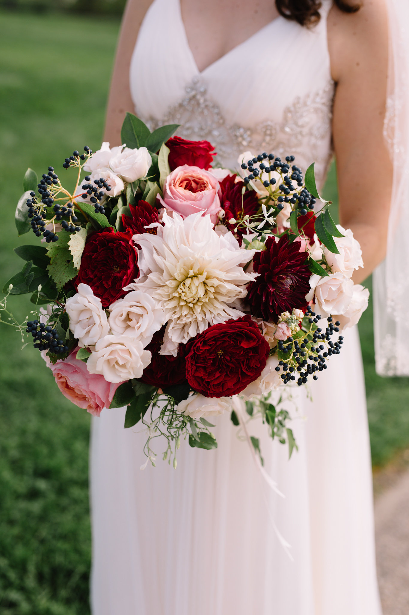 bride holding bouquet with dahlias, garden roses, berries, vines in red, coral, peach, blush