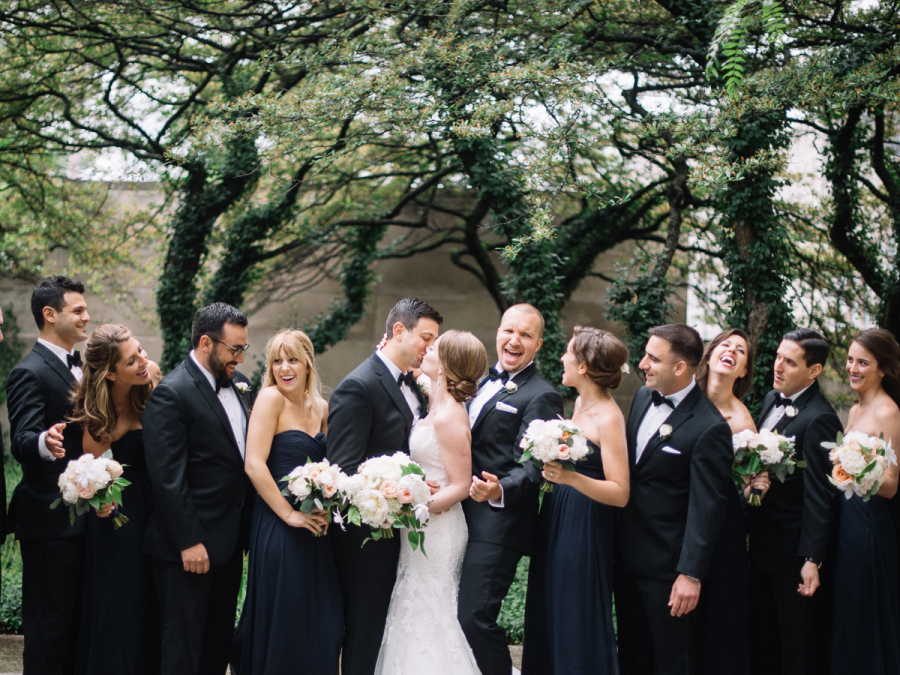 Wedding party in black, white, and navy, laughing with bouquets of white autumn clematis with vines, blush and ivory roses and peonies, and eucalyptus.