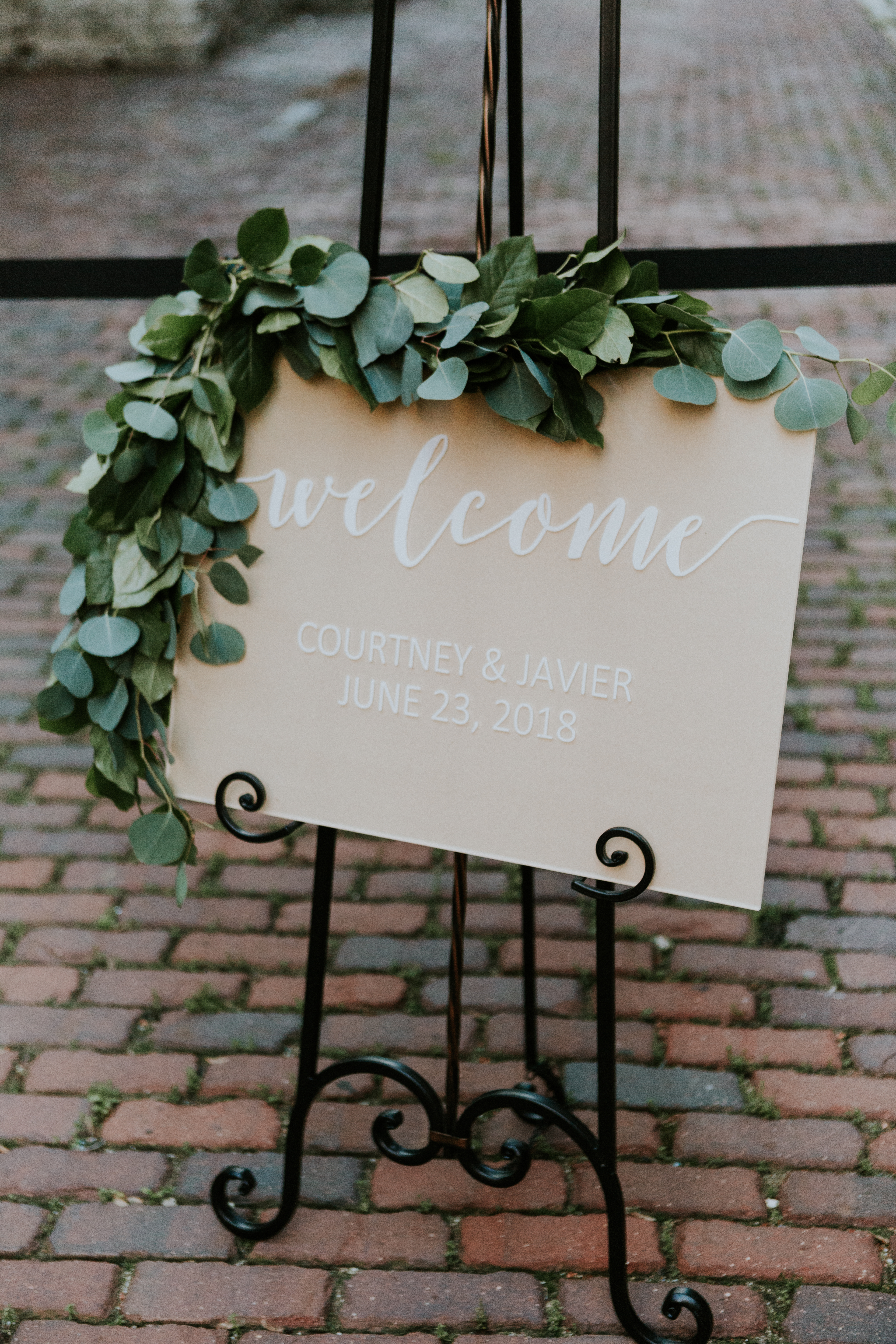 Simple blush welcome sign with eucalyptus garland on iron stand for summer wedding.