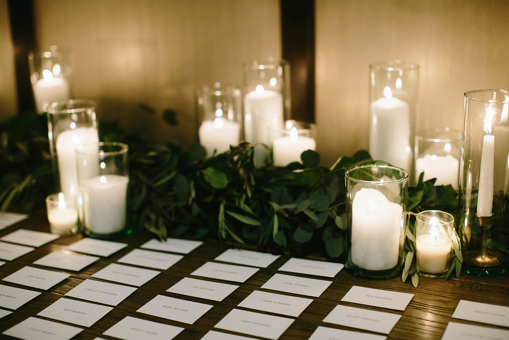 Placecard table at a winter wedding reception with white pillar candles in hurricane glass with olive foliage.