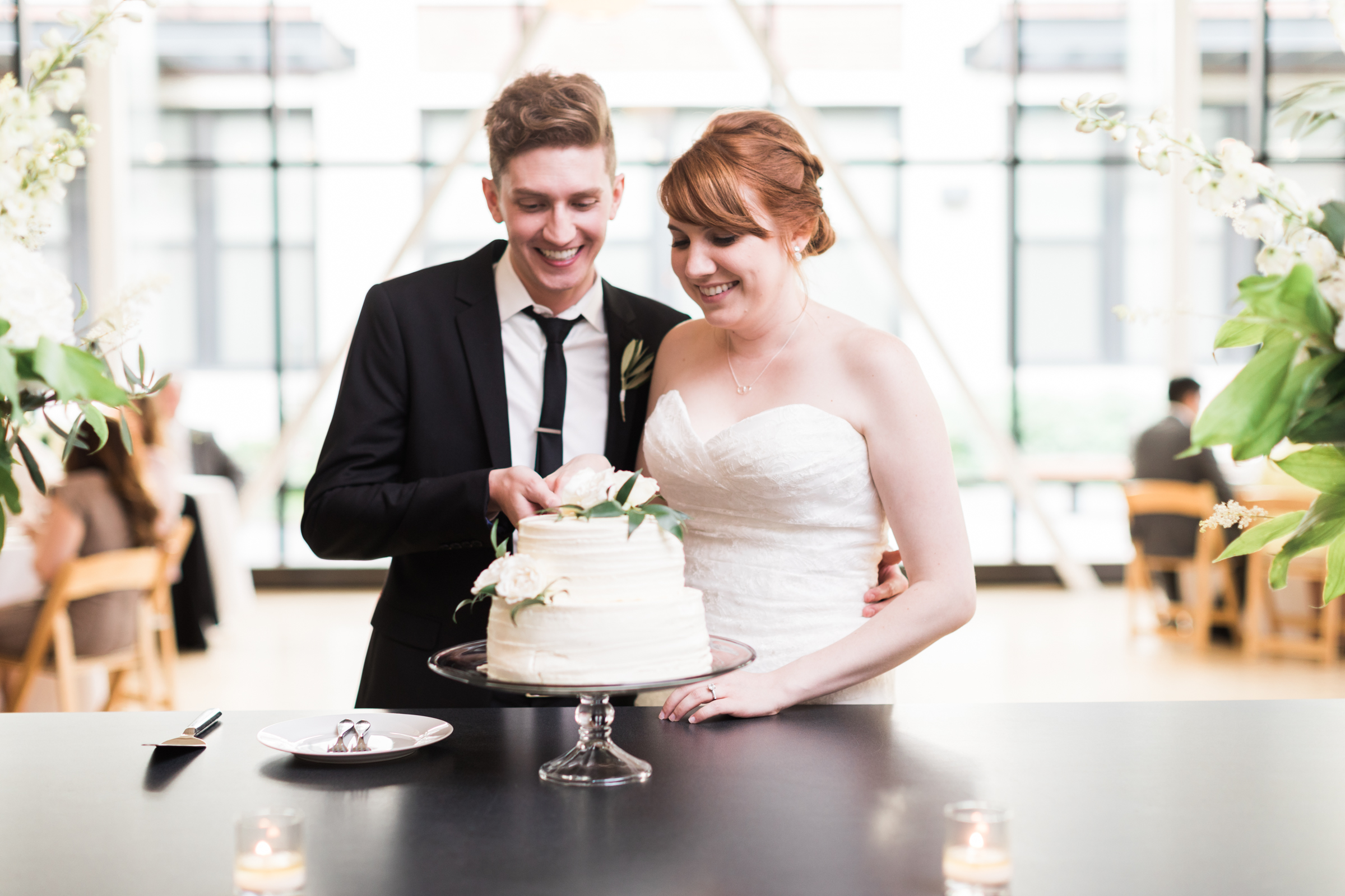 Bride and groom at Greenhouse Loft cut two-tiered white wedding cake with ranunculus for spring minimalistic wedding.