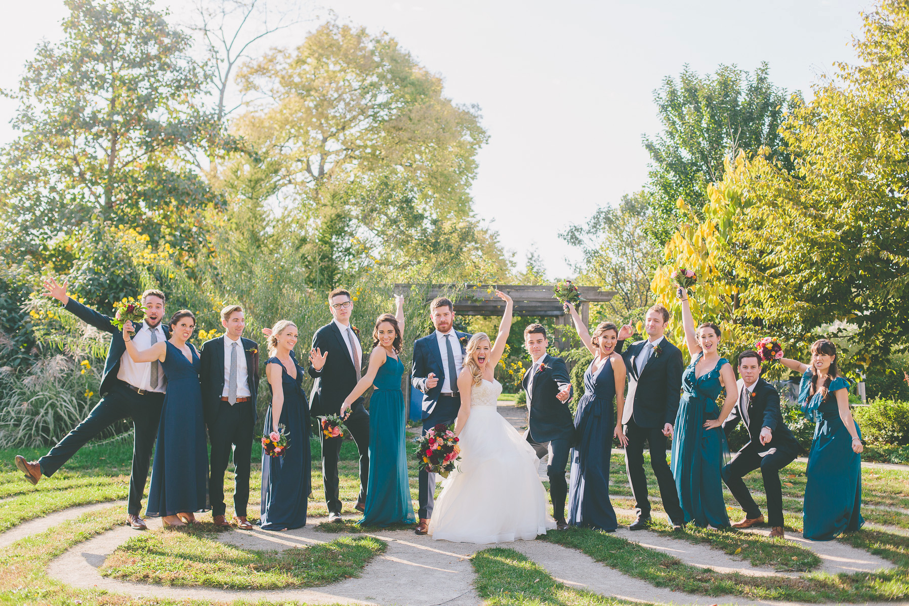Fall wedding party outside with teal dresses, orange ranunculus boutonnières, and bouquet with red, pink, plum, and bright yellow.