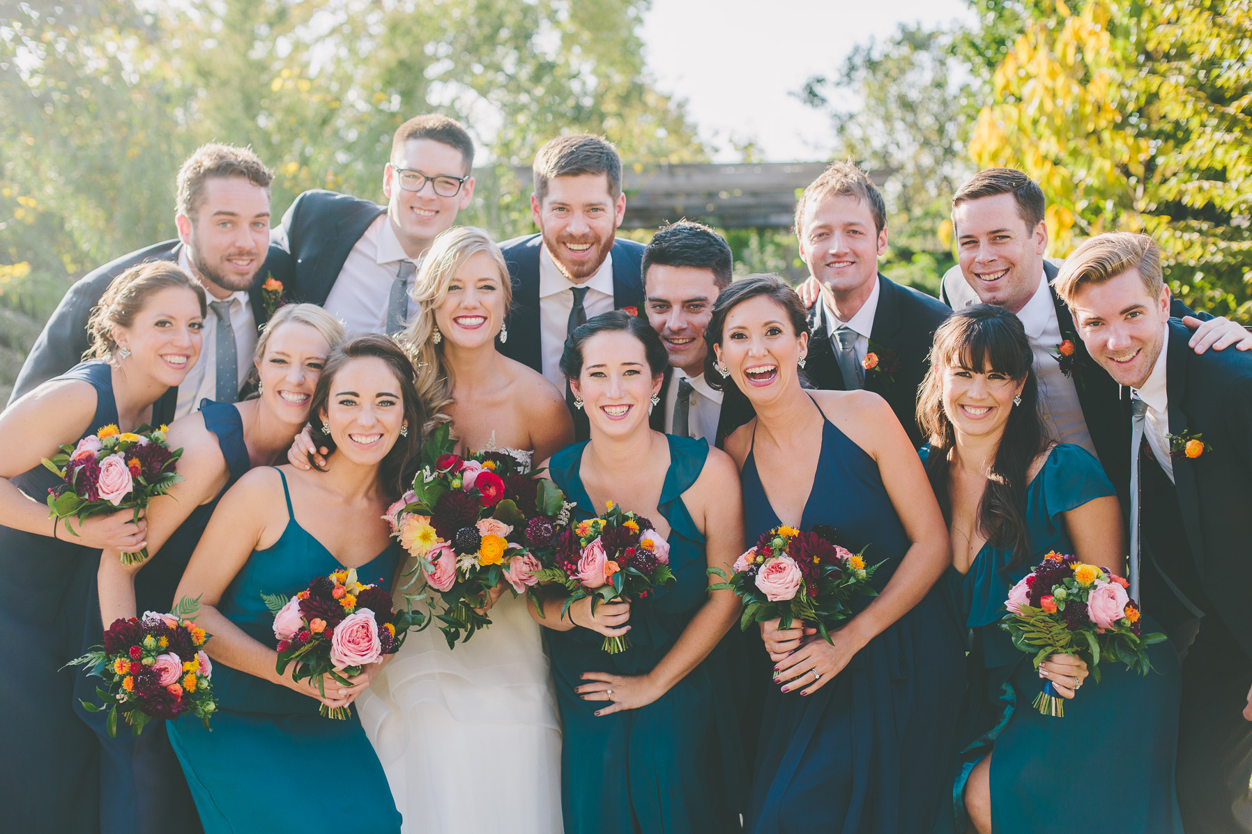 Fall wedding party outside with teal dresses, orange ranunculus boutonnières, and bouquet with red, pink, plum, and bright yellow.