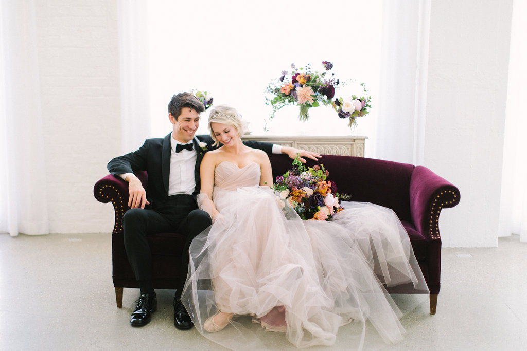 Groom in bowtie and bride in champagne dress with gardeny bouquet of pink dahlia and peonies, irises, plum scabiosa, and lilac on velvet couch..