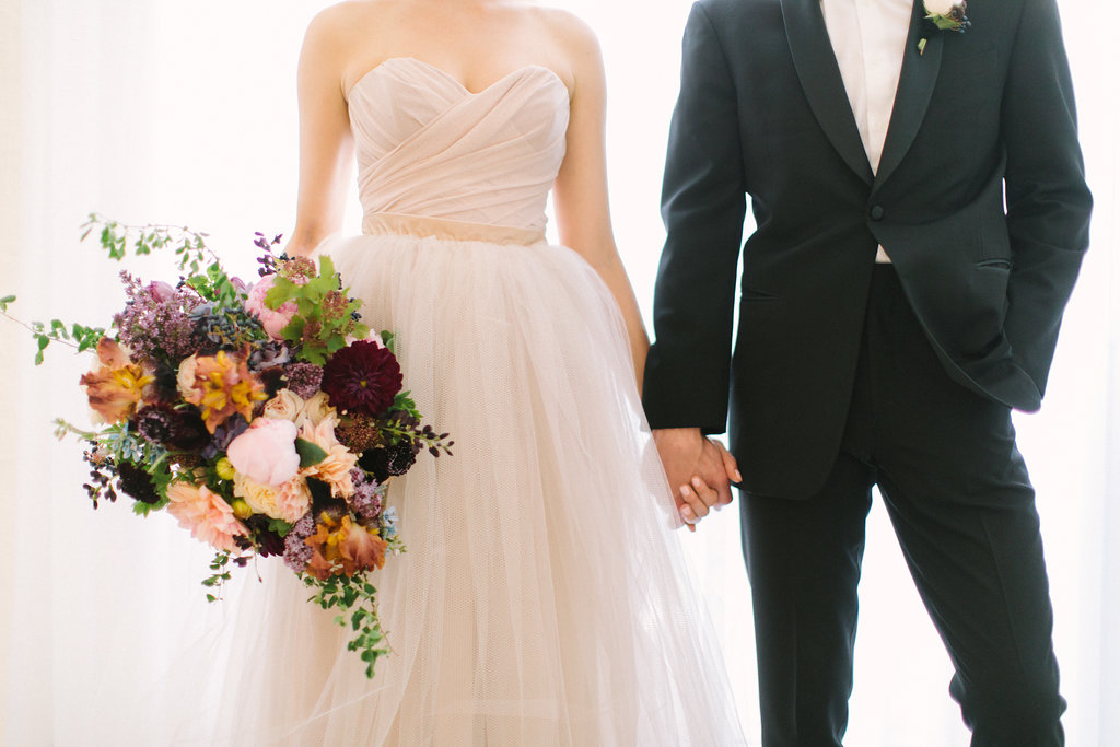 Groom in bowtie and bride in champagne dress with gardeny bouquet of pink dahlia and peonies, irises, plum scabiosa, purple hydrangea, berries, tweedia, and lilac.
