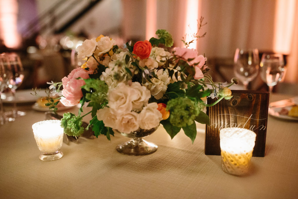 Orange ranunculus, pink peonies, white hydrangea, ivory roses, and snowball viburnum in mercury glass vase with tealight candles for spring wedding reception at Moonlight Studios.