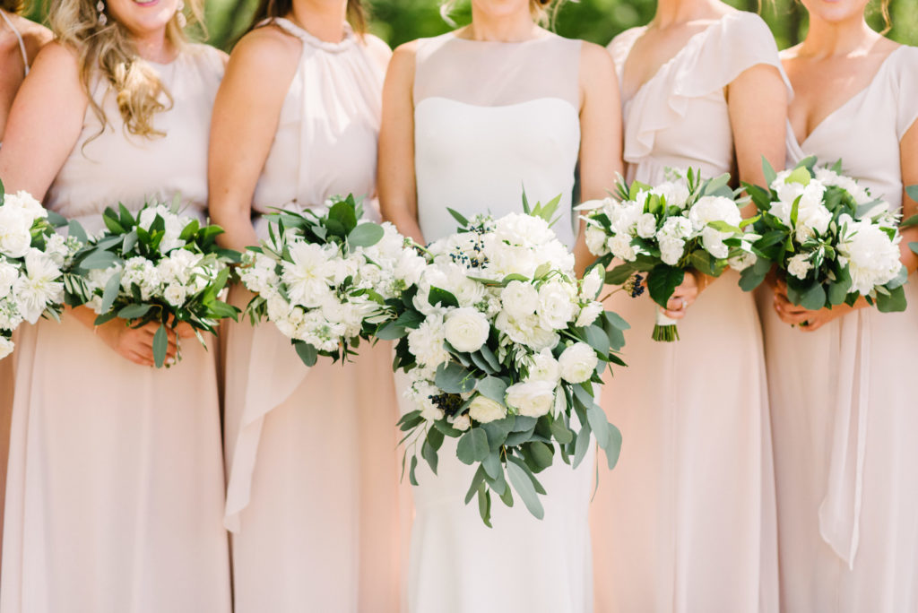 Bride and bridesmaids with white monochromatic bouquets of peonies, roses, and ranunculus with sage foliage.