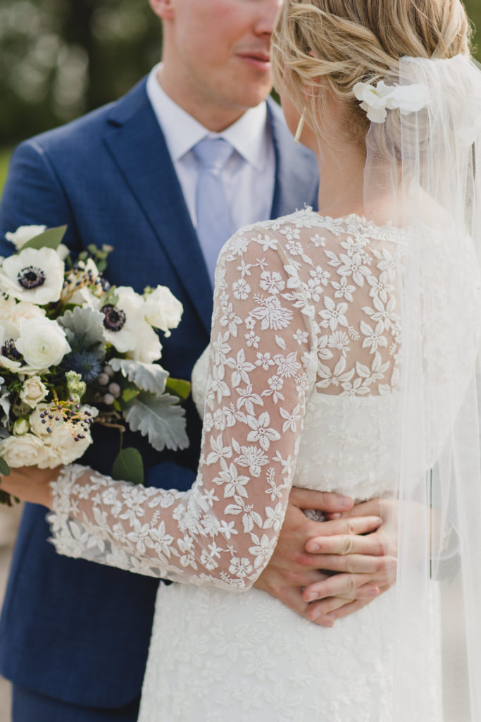 Bride and groom portrait outside; Bride wore a floral lace dress, draping veil, and held a bouquet of white spray roses, anemones, navy berries, dusty miller, thistle and eucalyptus in a subdued, monochromatic palette.