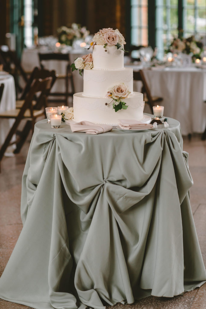 Three-tiered classic wedding cake with mauve and ivory dusty roses and japanese anemone on a sage green cake table with votive candles for a romantic outdoor wedding. 