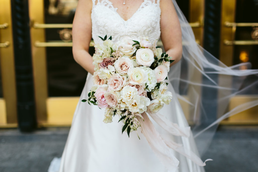 Bride with veil holding a classic bridal bouquet in ivory and blush with peonies, ranunculus, garden roses, and astilbe outside for her spring wedding.