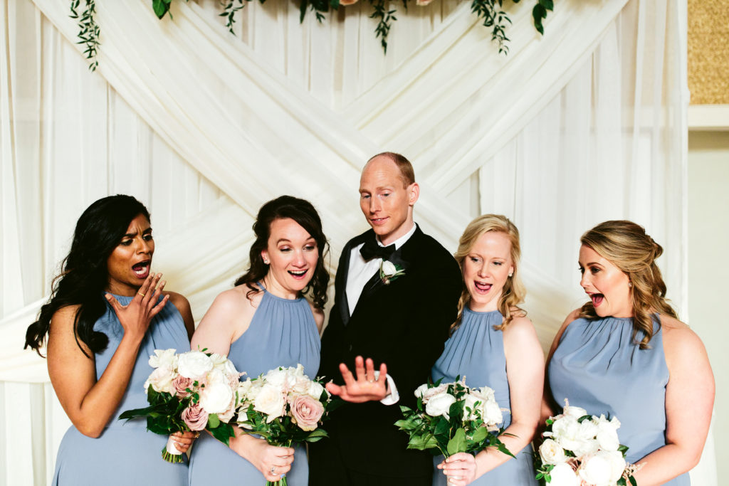 Bridemaids in dusty blue dresses holding bouquets of mauve garden roses, peonies, and ranunculus look at groom's ring in awe.
