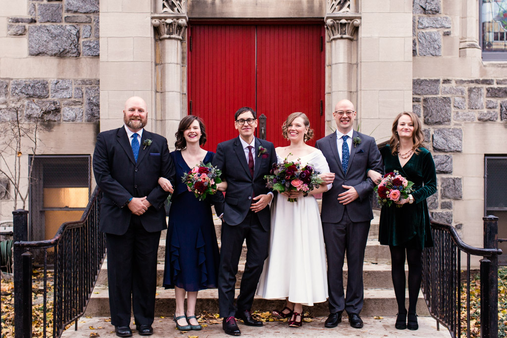 Wedding party smiles in front of red church doors for a fall ceremony featuring bouquets of pink garden roses, blue thistle, plum scabiosa, eucalyptus, fern, maroon dahlias, and berries.