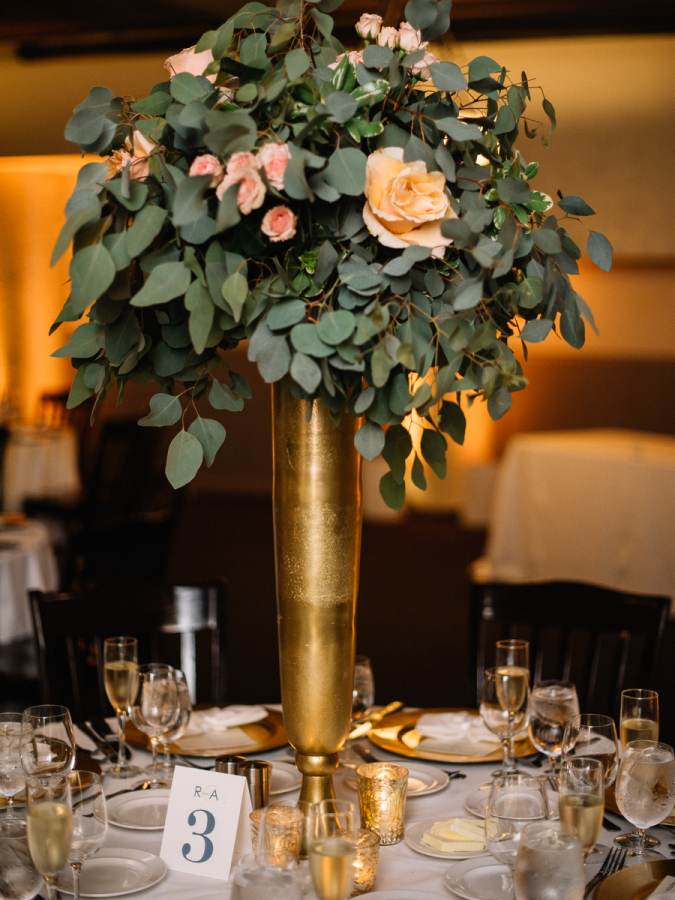 Elevated centerpiece with eucalyptus and roses in tall gold vase.