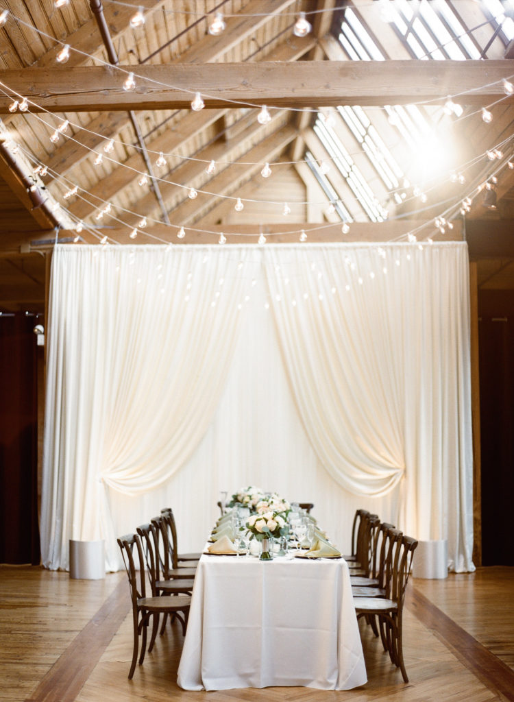 Bridgeport Art Center's Skyline Loft in Chicago wedding reception with elegant draping curtains, cafe lights, and head table in ivory with garden roses and eucalyptus. 