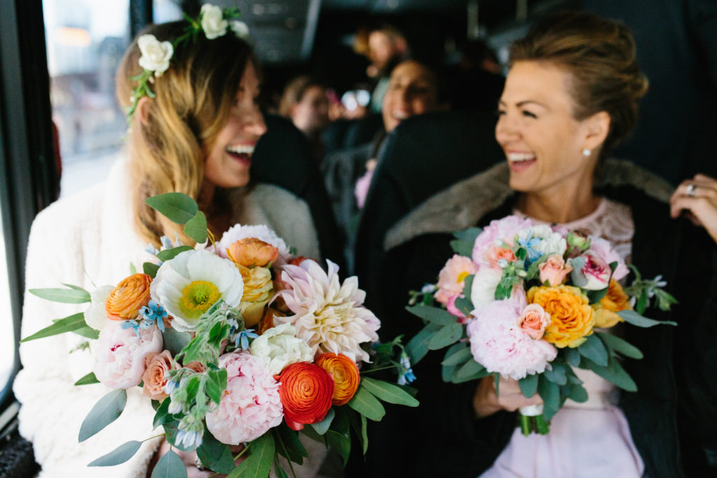 Bride and bridesmaid holding bright spring bouquets of orange garden roses and ranunculus, peonies, dahlias, dusty blue roses, and tweedia.