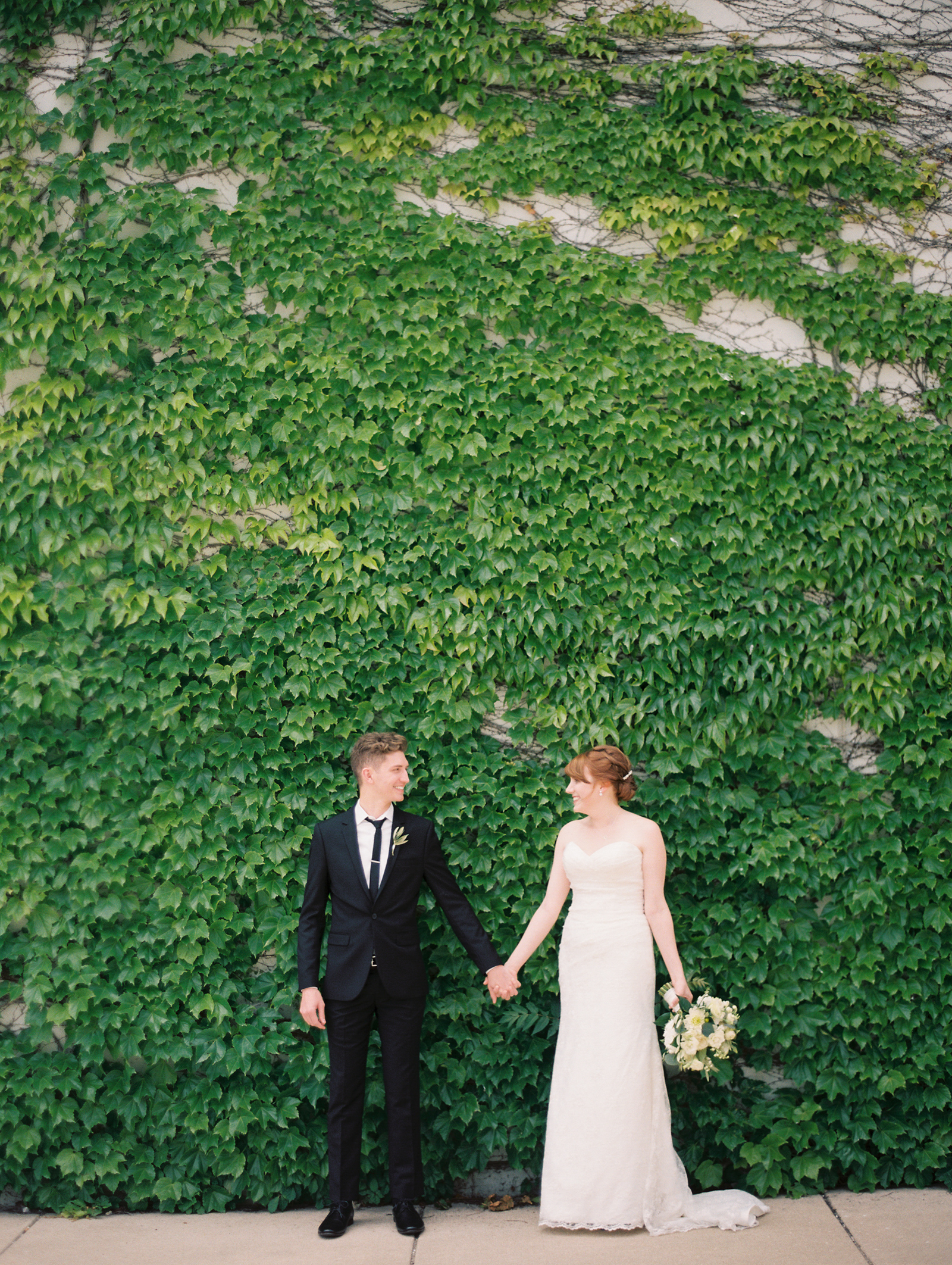 Bride and groom against vine covered wall with white bouquet of dahlias, garden roses, and eucalyptus.