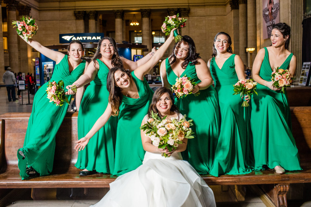 Spring bride and bridesmaid's in green dresses at Chicago Union Station with peach and pink bouquets of ranunculus, stock, and garden roses.