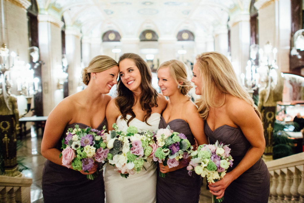 Smiling bride and bridesmaids with dusty purple and lime green bouquets of peonies, garden roses, snowball viburnum, anemones, and scabiosa. 