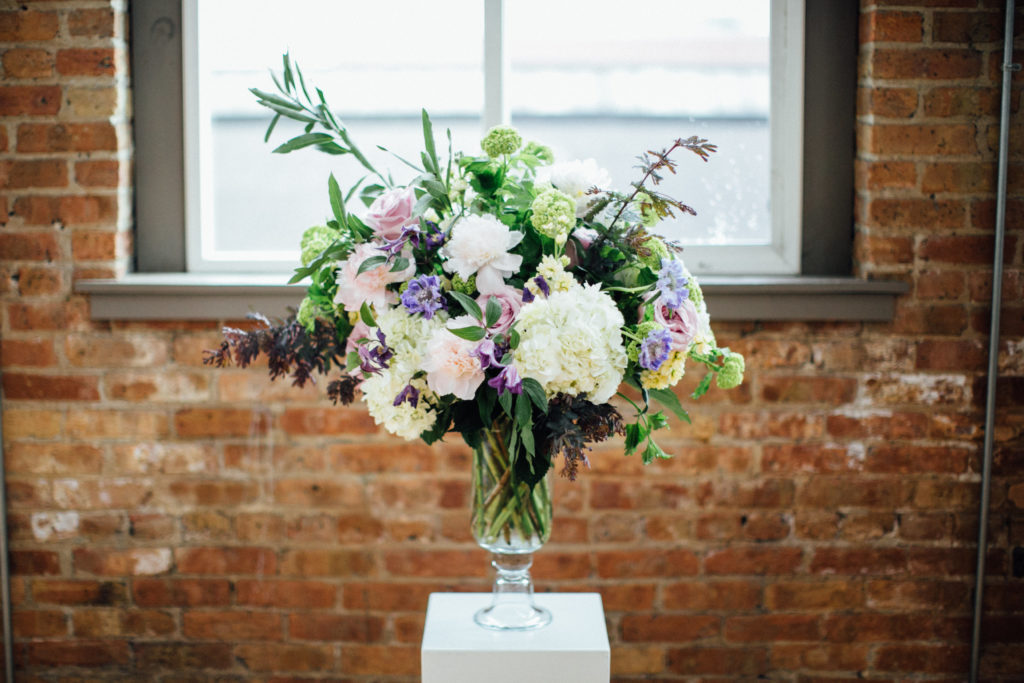 Main centerpieces in glass vases for spring wedding in purple and green, featuring peonies, hydrangea, scabiosa, clematis, and snowball viburnum. 
