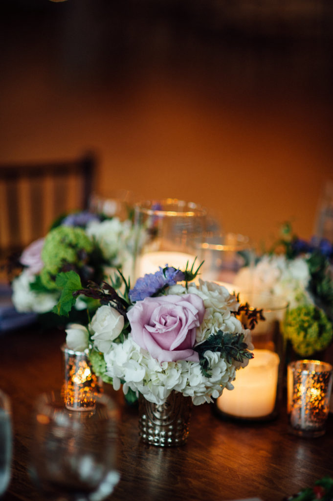 Pillar candles and mercury glass arrangements of garden roses, snowball viburnum, clematis, scabiosa, and hydrangea for spring wedding reception.