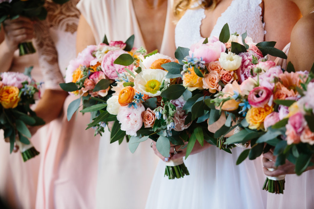 Spring wedding bride and bridesmaids with blush dresses holding colorful bouquets of pale pink peonies, orange ranunculus, tweedia, dusty blush spray roses and garden roses.
