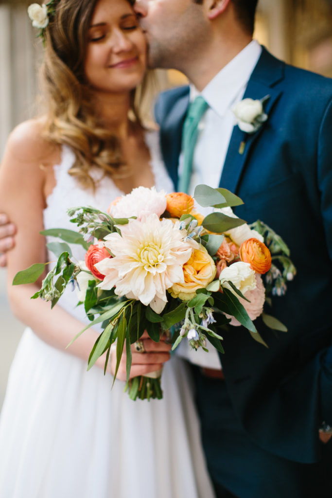 Smiling bride and groom with flower crown and colorful spring bouquet of orange ranunculus, peonies, ivory dahlias, eucalyptus, and blue tweedia.