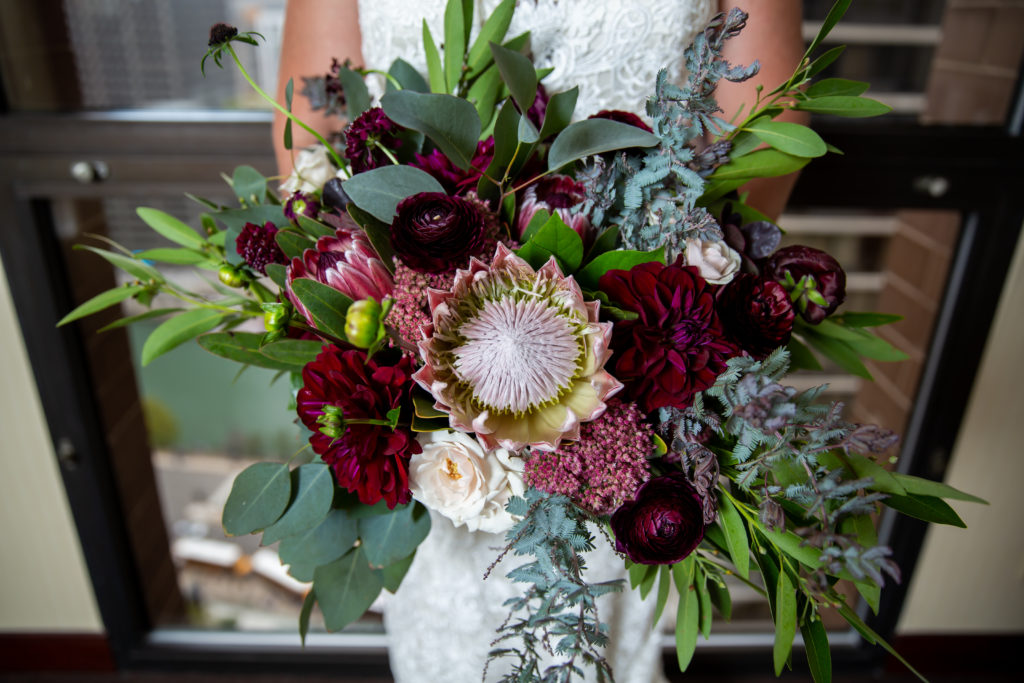 Jewel-toned bridal bouquet with protea, garden roses, scabiosa, purple acacia and white majolika for a wedding at Revolution Brewing. 