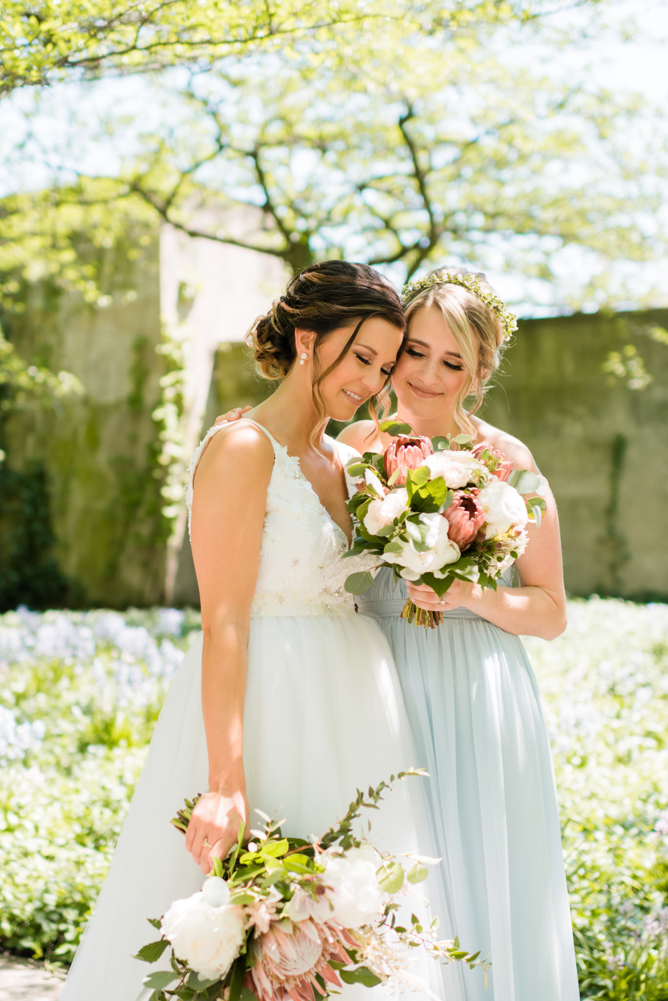 Bride and bridesmaid in garden for spring wedding, with bouquets of blush protea, white anemone, pale pink astilbe, and ivory peonies.