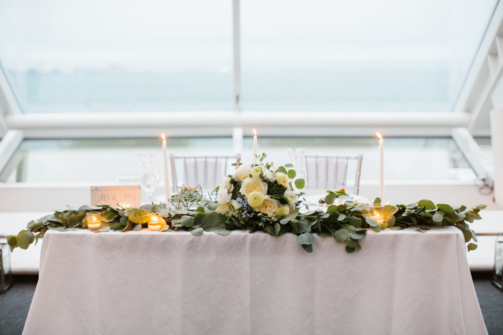Romantic sweetheart table at summer wedding at Adler Planetarium with white taper candles, eucalyptus swags and bridal bouquet of garden roses, ranunculus, tweedia, and succulents.