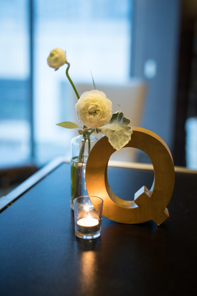 Simple details at spring wedding reception with ivory ranunculus and dusty miller, tea light candle, and golden 'Q' decoration. 
