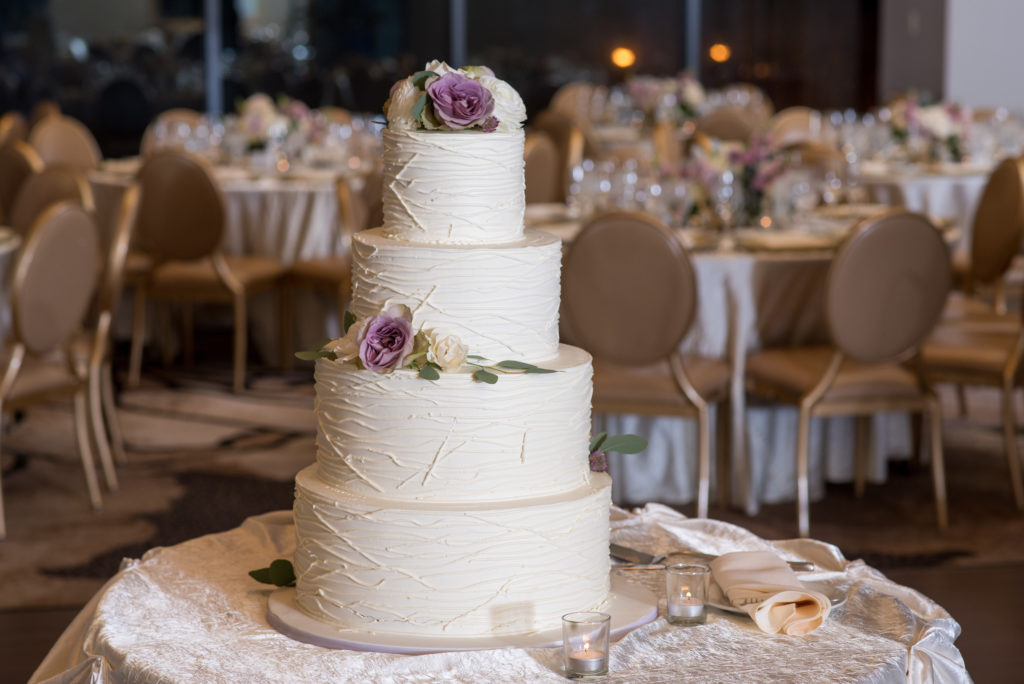 Textured four-tier spring wedding cake with ivory frosting and dusty purple garden roses.