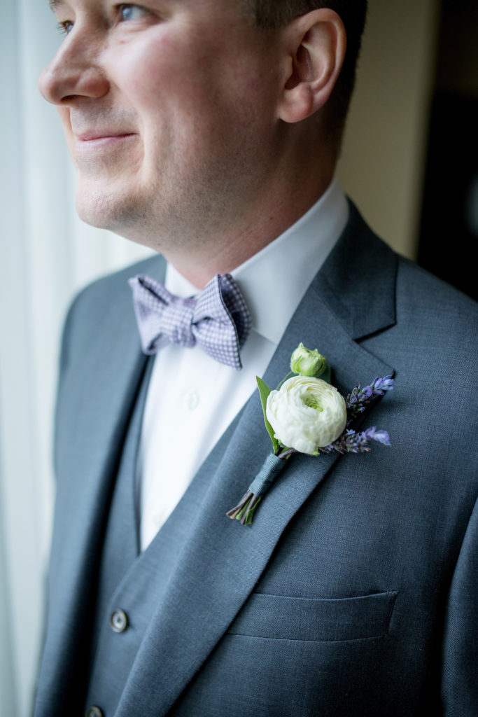 Spring wedding groom wearing a dusty purple bow tie and an elegant boutonniere of lavender and ivory ranunculus.
