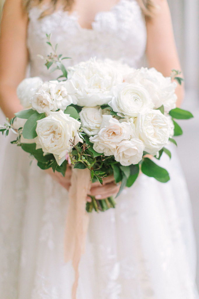 Bridal bouquet close-up with blush ribbon and ivory ranunculus, garden roses, peonies, and majolica roses for summer outdoor wedding.