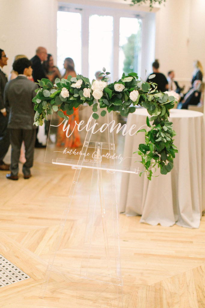 Clear acrylic wedding welcome sign for summer reception at Chicago Illuminating Company, with ivory garden roses and draping foliage for a simple and romantic look.