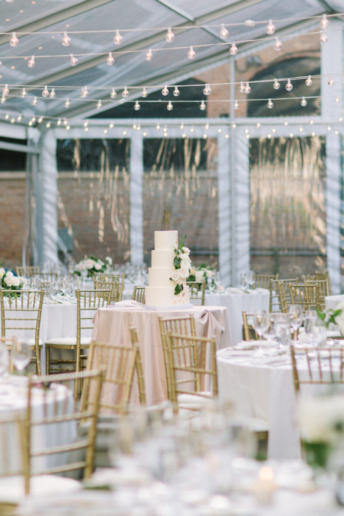 Romantic outdoor tented summer wedding reception in Chicago with four-tiered vanilla cake and cascading ivory roses with foliage, garden inspired arrangements, brushed gold bamboo chairs, and globe string lights. 