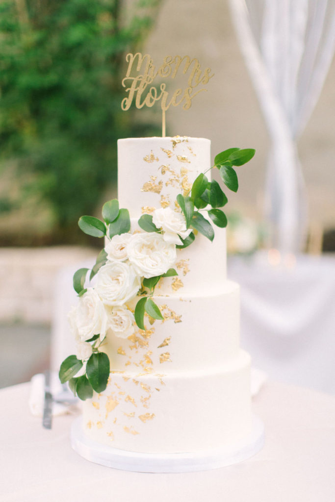 Four-tiered classic vanilla wedding cake with gilded gold flakes, cascading peonies, roses, and foliage with golden cake topper.