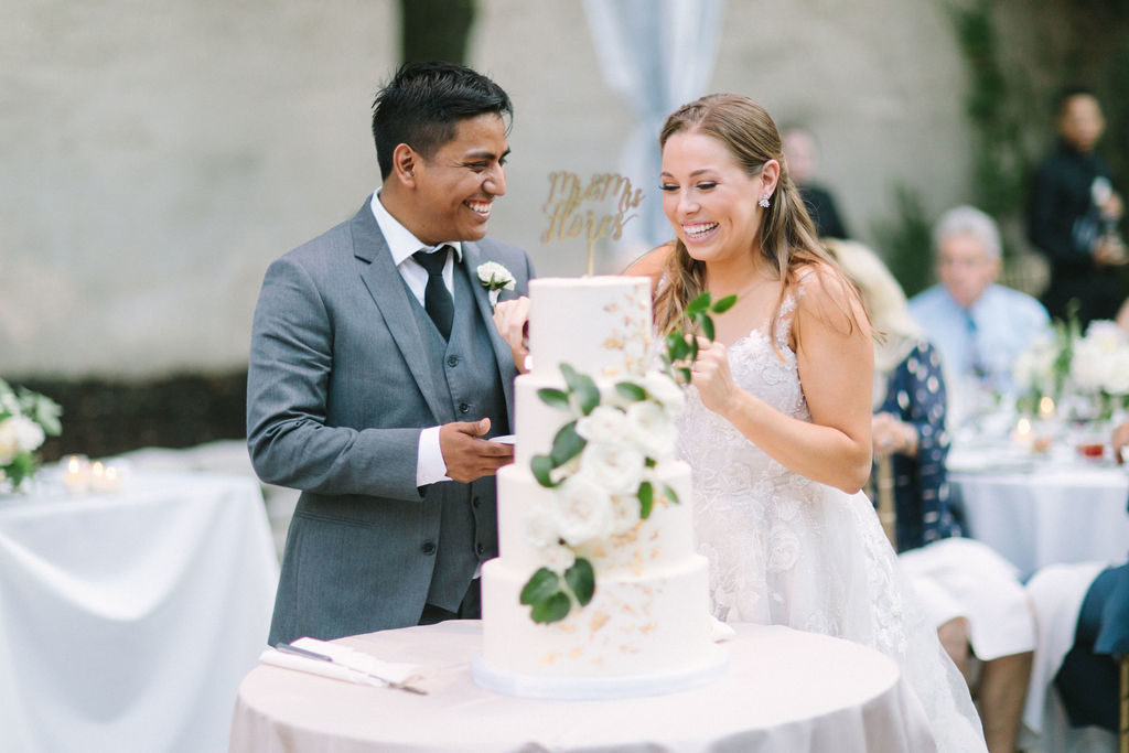 Smiling couple with a four-tiered classic vanilla wedding cake with gilded gold flakes, cascading peonies, roses, and foliage with a golden cake topper.