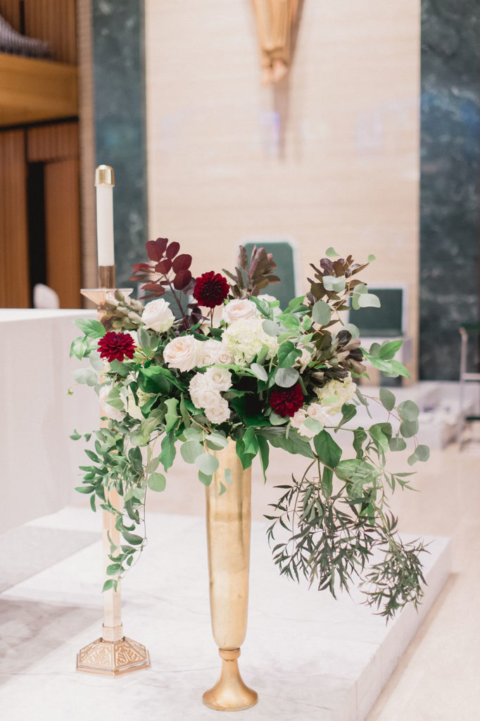 Tall golden vase for a fall wedding church ceremony with blush garden roses, pale green hydrangea, eucalyptus, burgundy dahlias, and spray roses, and draping foliage.