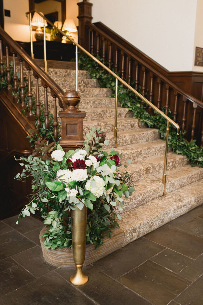 Romantic and dramatic tall brushed gold vase with a full arrangement of burgundy dahlias, hydrangeas, ivory garden roses, and foliage for a traditional fall wedding. 