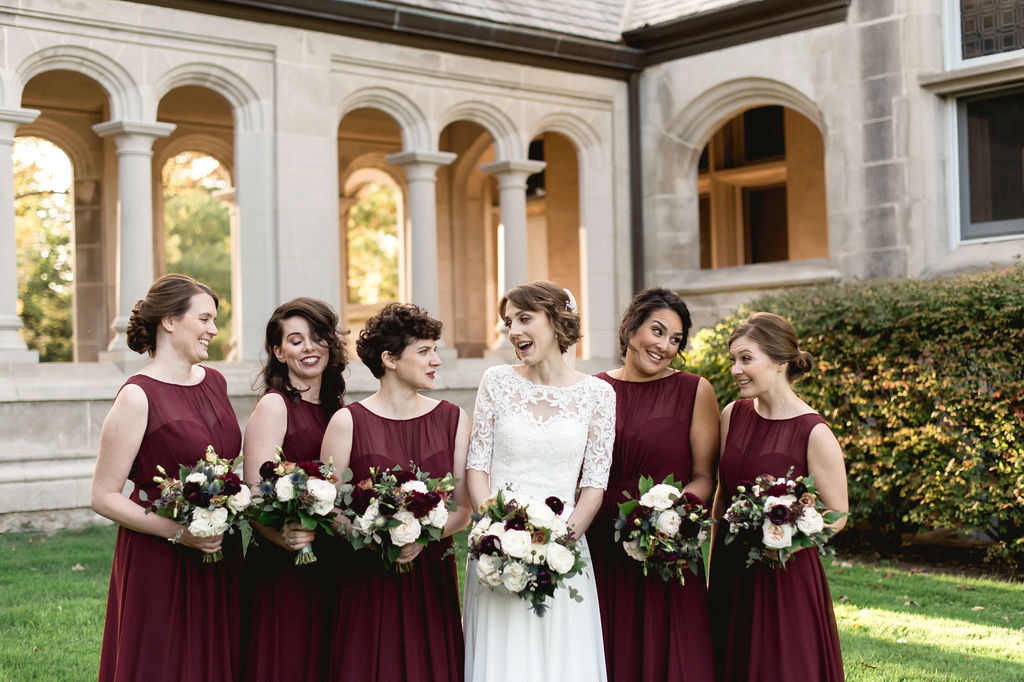 Bride and bridesmaids for fall wedding with burgundry dresses that match the bouquet's dahlias. Other flowers include plum ranunculus, ivory roses, thistle, berries, and dusty rose lisianthus. 