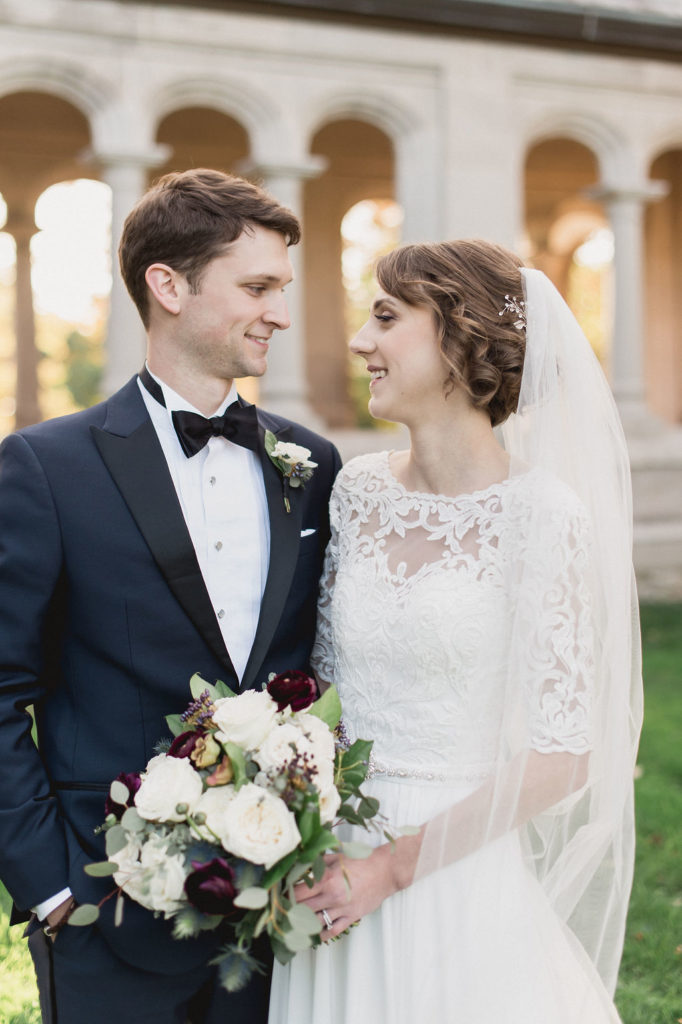 Bride and groom with a classic bouquet of plum ranunculus, ivory garden roses, dusty rose lisianthus, berries, and sage eucalyptus for their traditional fall wedding. 