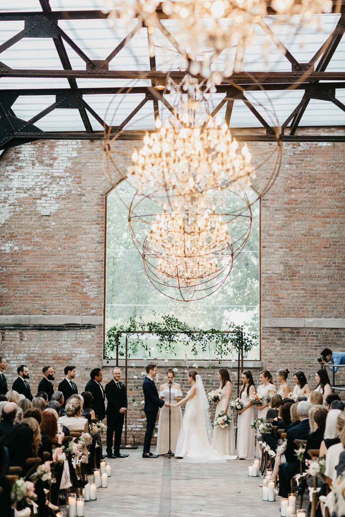 An elegant late summer wedding ceremony reception in Bridgeport Art Center's Sculpture Garden, featuring a chuppah with a hanging flower installation of ranunculus and stock. 
