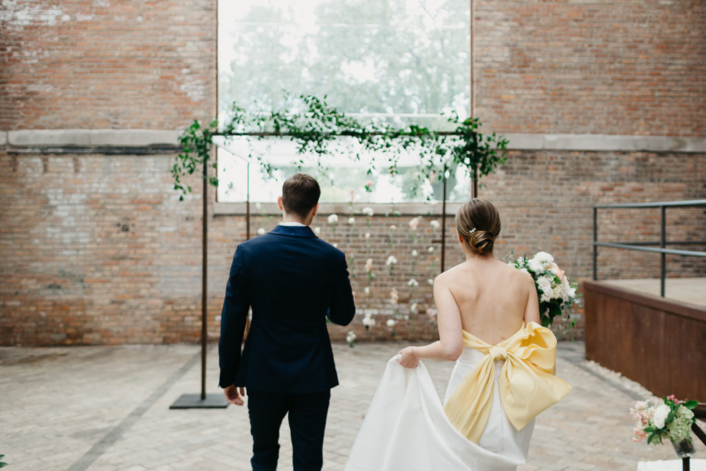 An elegant late summer wedding ceremony reception in Bridgeport Art Center's Sculpture Garden, featuring a chuppah with a hanging flower installation of ranunculus and stock. 
