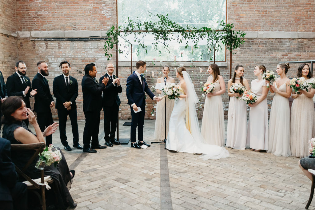 An elegant late summer wedding ceremony reception in Bridgeport Art Center's Sculpture Garden, featuring an industrial chuppah with a hanging flower installation of ranunculus and stock. 