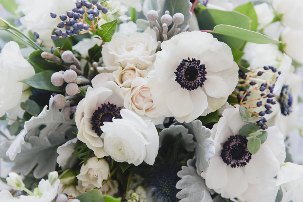Bridal wedding bouquet close-up with a subdued palette of white spray roses, anemones, navy berries, dusty miller, thistle and eucalyptus. 