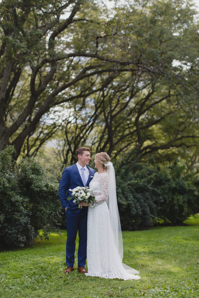 Bride and groom portrait outside; Bride wore a lace dress, draping veil, and held a bouquet of white spray roses, anemones, navy berries, dusty miller, thistle and eucalyptus; Groom wore a periwinkle tie and navy suit. 