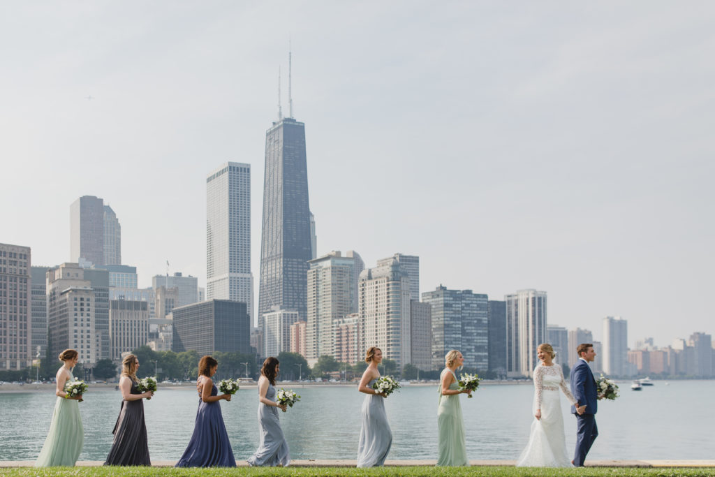 Bride, groom, and bridesmaids walking in a line in front of Chicago's skyline. The wedding palette of sage green, white, and dusty blue included flowers of anemones, spray roses, berries, thistle, dusty miller, and eucalyptus.