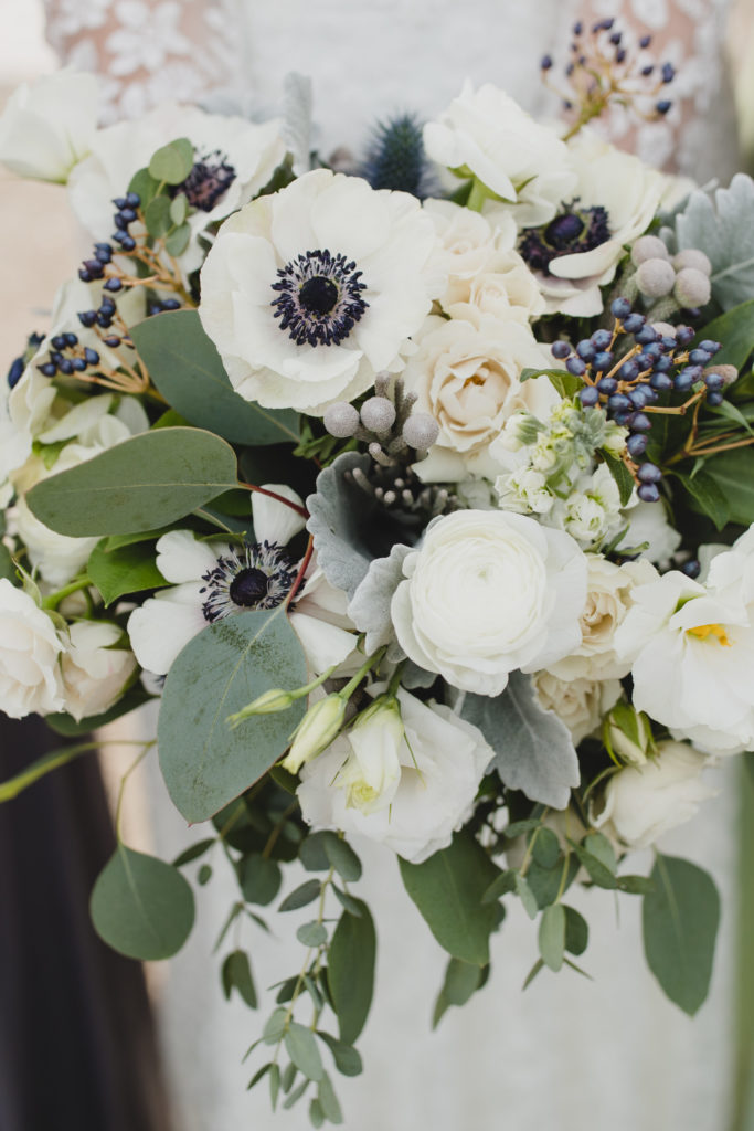 Mostly monochromatic navy, white, and sage green wedding bouquet close-up with a subdued palette of white spray roses, anemones, navy berries, dusty miller, thistle and eucalyptus. 