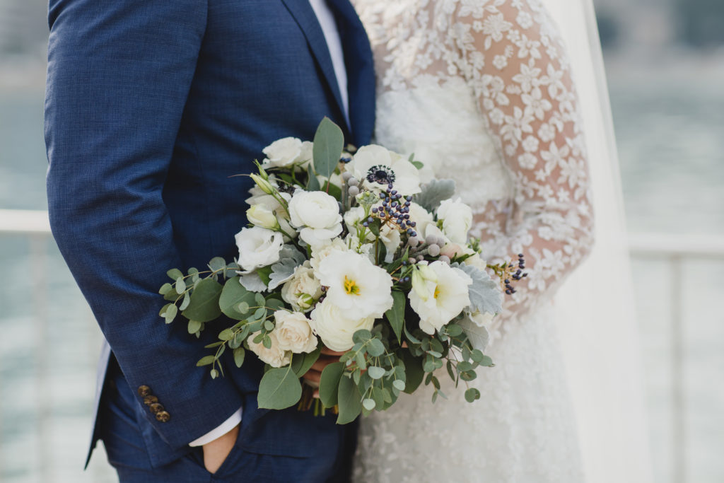 Bride and groom portrait outside; Bride wore a lace dress, draping veil, and held a bouquet of white spray roses, anemones, navy berries, dusty miller, thistle and eucalyptus in a subdued, monochromatic palette.